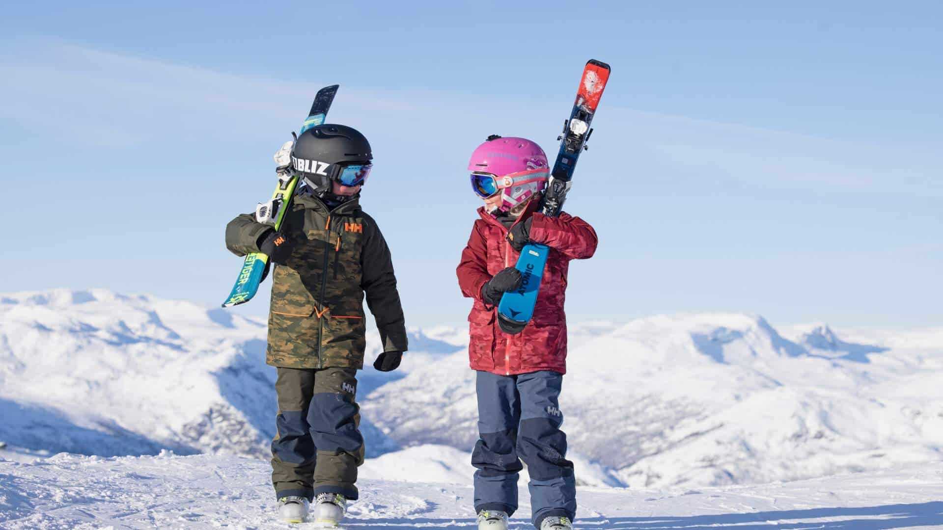 Two children in ski gear carrying their skis, mountains behind