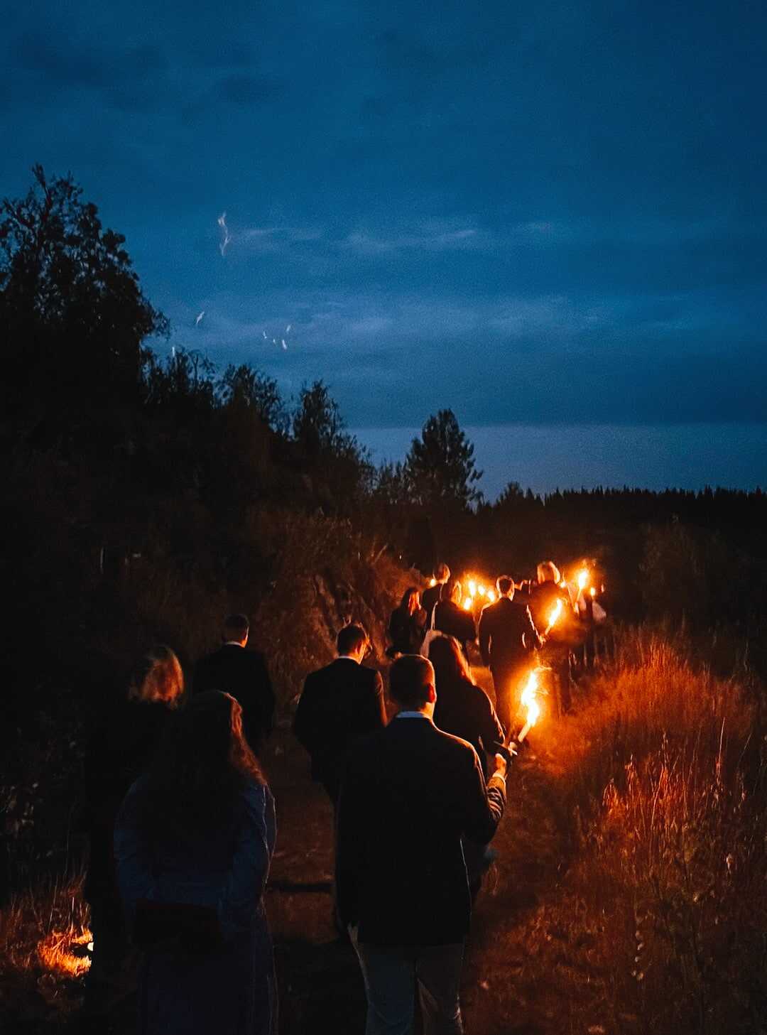 Wedding guests walk with torches along a forest path at dusk